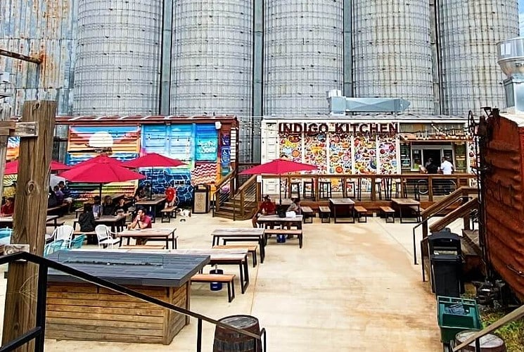 Food trucks and seating with red umbrellas in an open yard in front of several large silos