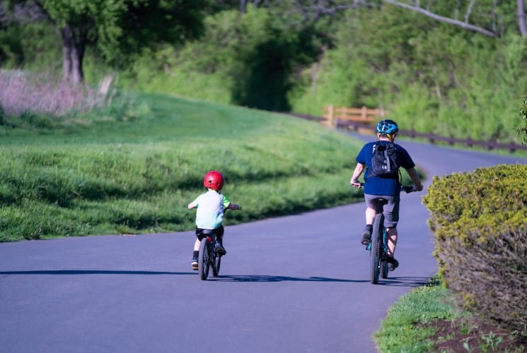A man and boy riding bikes on a paved path surrounded by lawn and trees