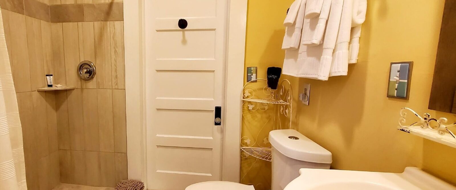 Bathroom with tiled stand up shower, pedestal sink, toilet, yellow walls and white towels hanging on a rack