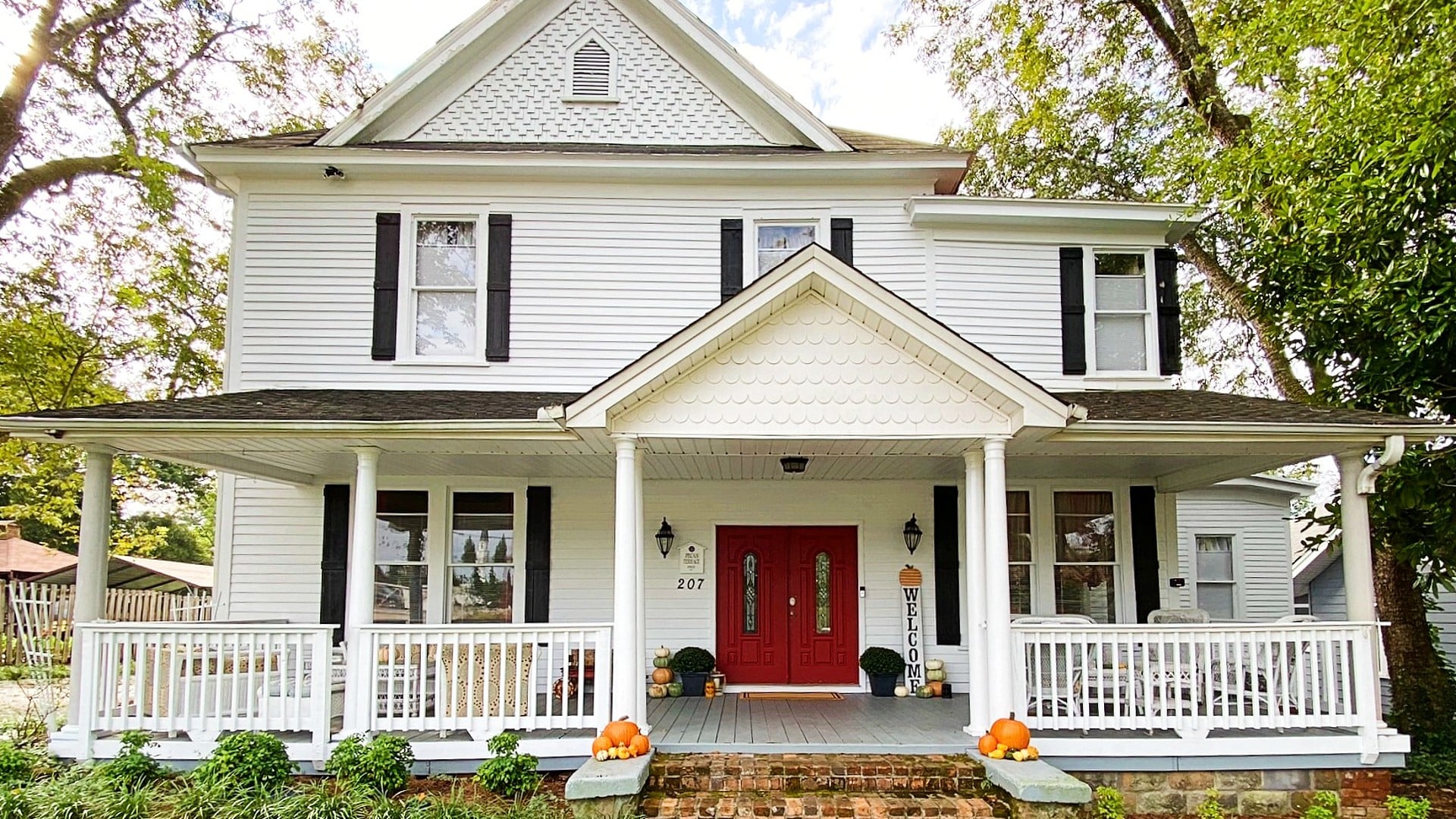 Front facade of a white two-story home with large front porch, black shutters and a red door.