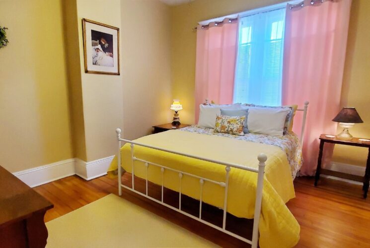 Quaint bedroom with white wrought iron headboard and footboard, yellow walls and window with pink curtains