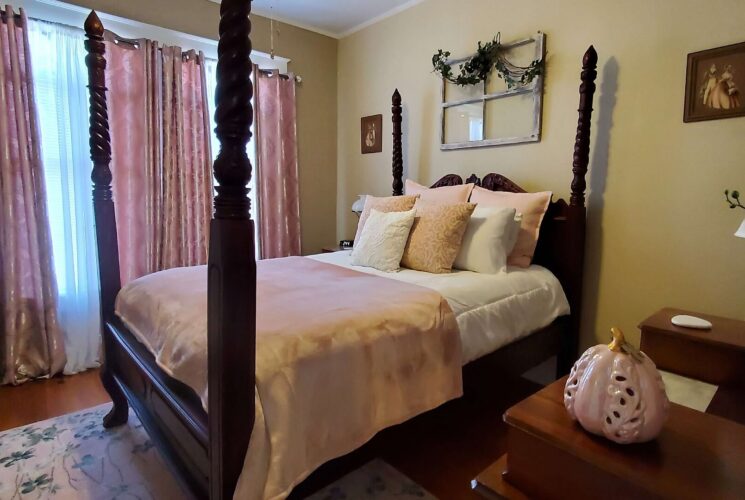 Elegant bedroom with four poster mahogany bed in pink and white linens with an antique dresser and large windows with curtains