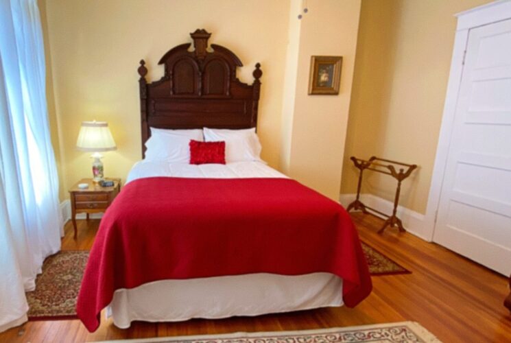 Elegant bedroom with queen bed with tall antique headboard, hardwood floors and bright windows with white curtains