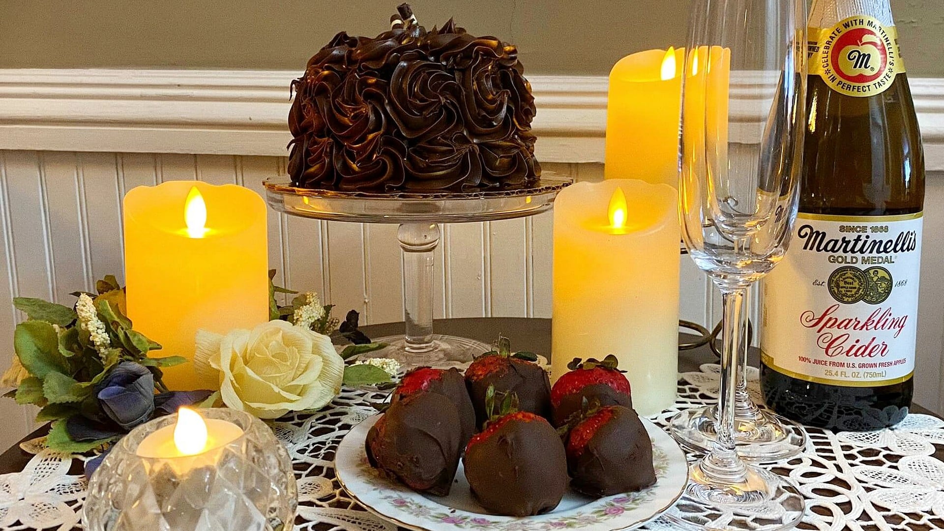 Table set with candles, chocolate cake, chocolate dipped strawberries, and bottle of cider with two flutes