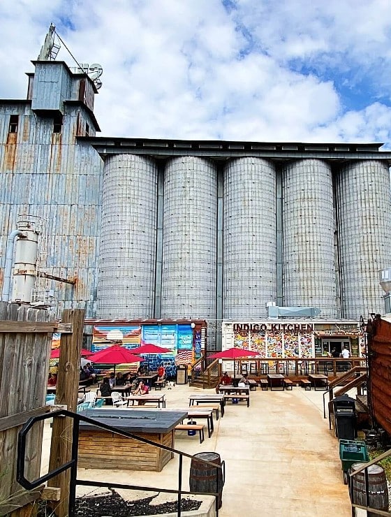 Food trucks and seating with red umbrellas in an open yard in front of several large silos