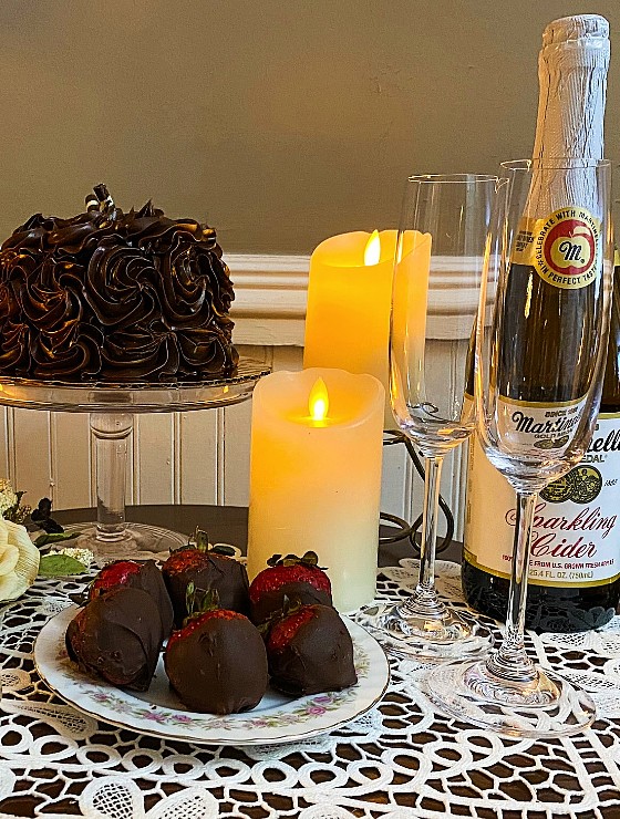 Table set with candles, chocolate cake, chocolate dipped strawberries, and bottle of cider with two flutes