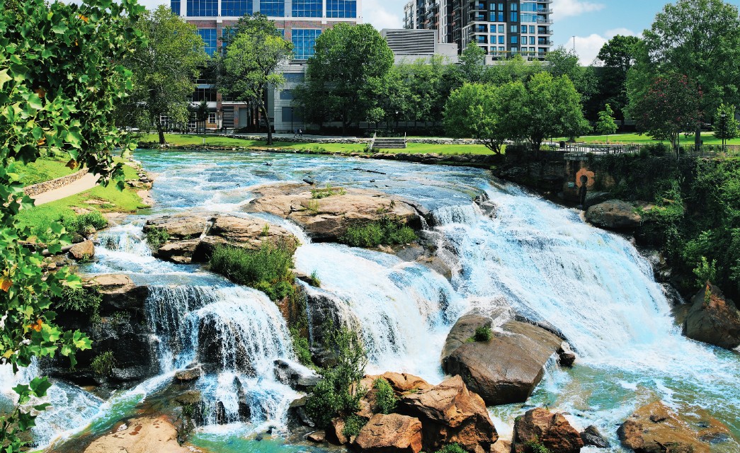 A cascading waterfall from a small river by grass and trees next to city buildings